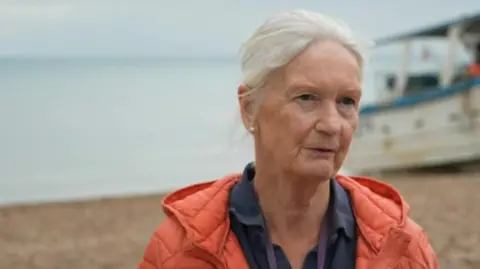 A white-haired lady in an orange jacket and blue shirt is on a pebbly beach with an out of focus boat in the background. 