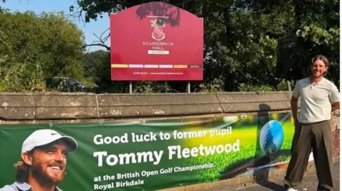 Scarisbrick Hall School Tommy Fleetwood stands outside in front of a sign reading: 'Scarisbrick Hall School'. Underneath is a banner reading: 'Good luck to former pupil Tommy Fleetwood at the British Open Golf Championship Royal Birkdale.'
