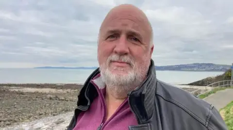 David Graves. He has a bald head and short grey beard. He is wearing a black jacket, perhaps made from leather, with a purple zip up top underneath