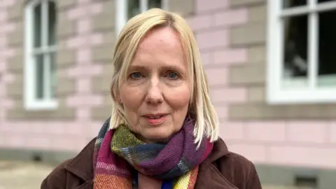 Catherine is standing outdoors in front of a pale pink building with white-framed windows. She is wearing a dark coat and a multicoloured scarf, with stone paving in the foreground.