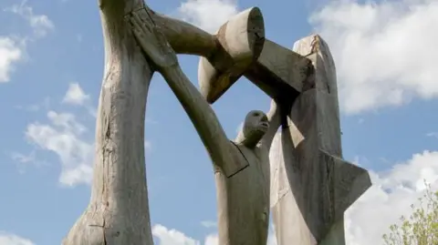 Close-up of the top section of the Peterborough Arch sculpture by Lee Grandjean. Behind it is blue sky and clouds. It shows an abstract wooden man with his arms up, touching the top of the wooden arch. 
