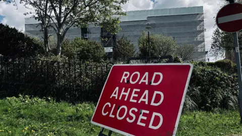 This image shows a roadside scene dominated by a large red temporary traffic sign in the foreground. The sign reads “ROAD AHEAD CLOSED” in bold white capital letters and is mounted on a black metal frame standing on a patch of grass. 