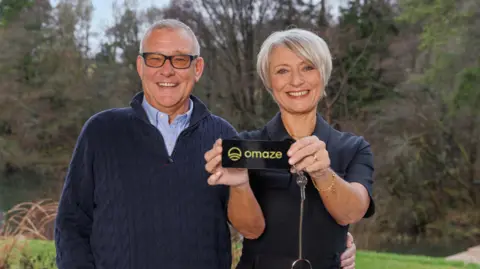 Omaze Martyn and Nicola Dickinson stand in front of a forested area. They are holding a key which says "Omaze". The man has his hand on the woman's waist and they are smiling widely.