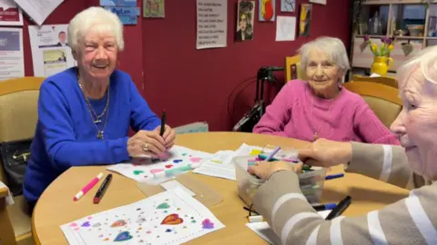 Three pensioners, Alice, Mary and Margaret sitting at a table with felt-tip pens and colouring sheets. They all have short, white or grey hair. Alice is wearing a blue jumper and gold necklaces, Mary is wearing a pink jumper and Margaret is wearing a brown and white striped jumper.