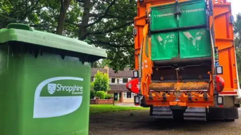 Shropshire Council A green bin with a Shropshire Council logo on it and a bin lorry in the background