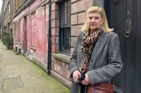 Julia Kuznecow wearing a black and white jacket and carrying a brown handbag standing outside a row of 18th Century houses