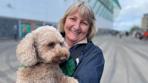 A woman in a blue jacket and green scarf holding up a dog in her arms towards the camera
