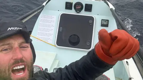 Tom Waddington wearing a black waterproof jacket and cap gives a thumbs up with his left hand while rowing across the Atlantic ocean.