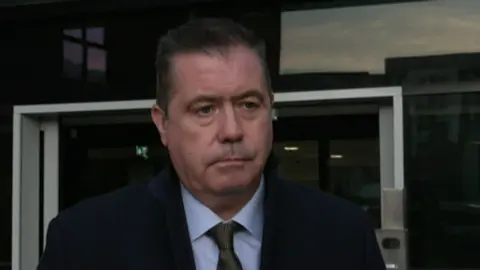 Glyn Roberts looks straight at the camera. He is wearing a black suit, grey tie and blue shirt. He has black/grey hair.