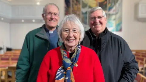 Three people stand inside a church with rows of chairs behind them. In the centre, Sue Gilbert, an elderly woman, smiles at the camera and wears glasses, bright red cardigan and blue scarf. Behind her stand two smiling men, one is wearing a clerical collar.