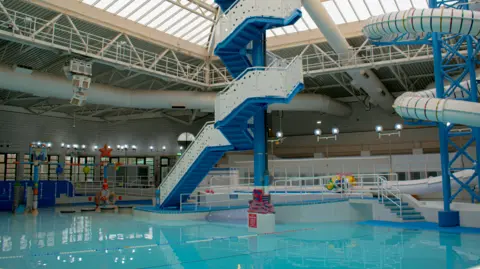 Leisure Focus An indoor swimming pool. There is a spiral tower of steps leading to a slide. The pool is aqua blue.