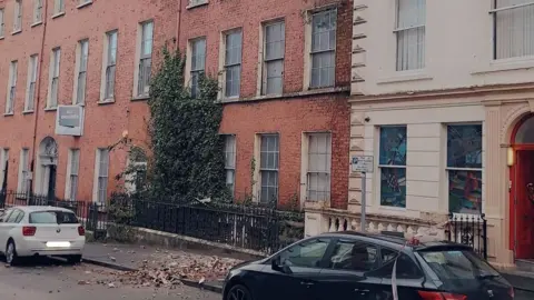 PSNI A street photo showing a row of terraced Georgian-style buildings. A pile of bricks lie on the pavement below the building in the middle of the photo. Cars are parked on either side of the bricks.