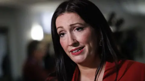 PA Media A close-up image of Emma Little-Pengelly. She has long, dark hair and is wearing a red suit jacket. She is mid-speech and looking at a TV reporter to her side.