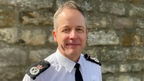 A man - Chief Constable Paul Gibson wearing a white shirt with a black tie, he has short grey hair and is standing in front of a grey stone wall.
