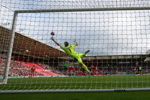 Tom Banks/MFC A goalkeeper in green dives to save a ball. The picture is taken from behind the net.