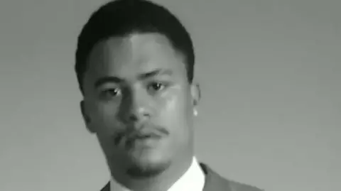 A black‑and‑white portrait photograph of a person wearing a suit jacket, white shirt and tie, facing the camera against a plain background.
