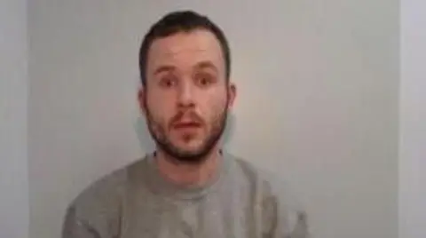 Greater Manchester Police A white man with a short beard, short brown hair and wearing a grey jumper looks at the camera