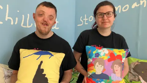 (l-r) Cameron Locke and Emma Stallard both looking at the camera are wearing black t-shirts and are each holding a pillow which they have made.