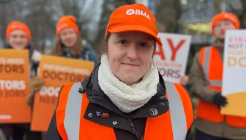 Sarah Sanderson/BBC Resident doctor Becky Lavelle standing on a picket line. She is wearing an orange hi-vis vest and an orange cap with the letters BMA on it. Behind are a group of other people, wearing orange hats and holding placards.
