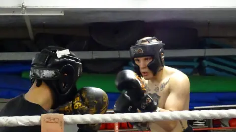 Daniel Bainger Daniel in a boxing match with one other person. They both are wearing helmets. Daniel does not have a top on, the other man is wearing a black T-shirt.