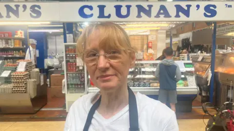 Photograph of Kathleen Leigh, who runs the Clunan's food stall at Wigan Indoor Market. She is pictured in front of her store, with customers in the background.