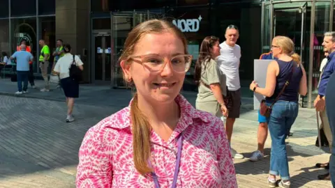 Heather Taylor with long blond hair in a ponytail wearing glasses and a pink and white shirt. She stands outside the Plaza building in Liverpool city centre. She is smiling. Protesters stand behind her.