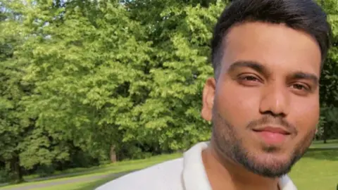 Mohammed Armaan  A person is standing in a grassy park with lush green trees in the background. The individual is wearing a short-sleeved white polo shirt. The lighting suggests a sunny day, and the image includes text overlay that reads “Armaan Wednesday” in white letters near the lower left side.