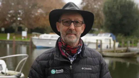 Jamie Niblock/BBC Man wears a fedora-type hat, red check scarf and black puffer-type jacket. Behind him are boats on a water course.