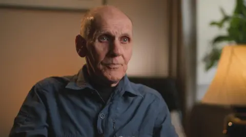 Ronnie Weir, an older man with thinning hair and wearing a dark blue shirt, is sitting in a living room.