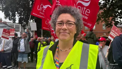 BBC/Crispin Rolfe Woman with curly hair, glasses, fluorescent bib and dark top stood in front of a crowd of people with unite the Union flags.