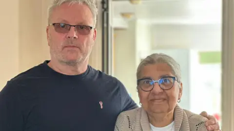 Jeff Dawes is seen in a dark t-shirt standing with his arm round Coleen, who is wearing glasses and a beige cardigan, both posing indoors and looking at the camera.