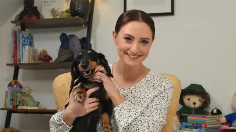 A woman with light brown/ginger hair slicked back with a middle parting looking at the camera and smiling. She is wearing a white top with black spots on. She is sitting in a yellow chair. She is holding a black and brown sausage dog who is licking its lips. In the background is a ladder shelf with books and toys.