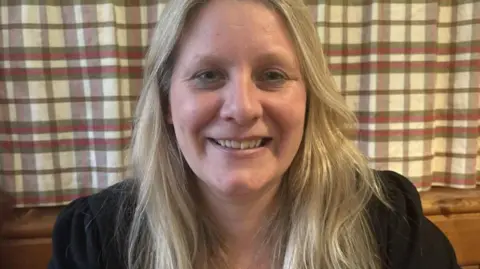 Sarah Evans pictured from the shoulders up. She has long blonde hair, is wearing a black jacket and is smiling at the camera. Behind her there is a red, green and cream, gingham curtain.