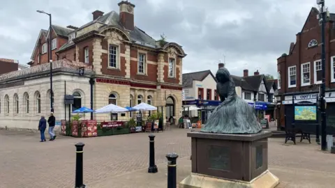 A town centre square. There is a statue in the middle of the square. It is of a woman sitting down with her dress swooping onto the floor and covering her feet. There are black bollards around it and shops and cafes can be seeing behind the statue. The centre is pedestrianised and two people are walking out of shot.