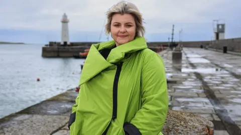 Tara Lynne O’Neill pictured at Donaghadee harbour with lighthouse in background, wearing a bright green jacket 