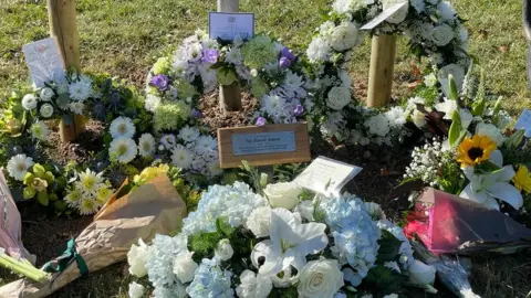 Stephen Huntley/BBC Floral tributes laid at the foot of a tree planted on the anniversary of Sir David Amess' murder