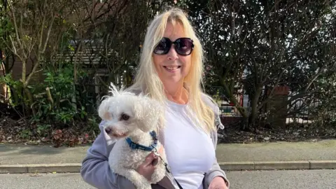 Sheena Stoddard Sheena, a blonde lady wearing sunglasses, is holding a small fluffy white dog, Henry, and smiling.