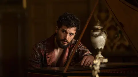 BBC/Bad Wolf Productions/HBO/Simon Ridgway Kit Harington as Henry Muck, wearing a silk dressing gown, leaning over a grand piano