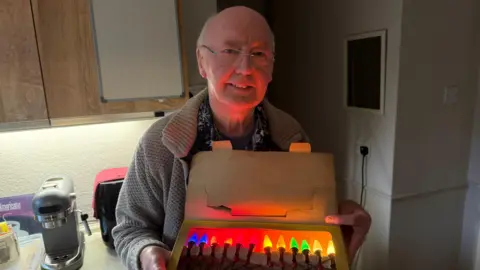 BBC / Grace McGrory The picture shows a man smiling at the camera. He is bald and is wearing glasses. He has a brown cardigan on, and a blue patterned shirt. He's standing in a kitchen. Behind him you can see a kitchen counter and cupboards. He's holding an old cardboard box, which is filled with a string of multi-coloured lights. 