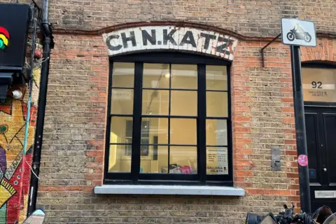 Black framed window in an exterior brick wall with a brick archway above it and CH N.Katz written in black over it