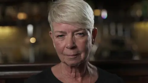 Head and shoulders shot of a woman looking at the camera. She has short blonde hair, grey-blue eyes and is wearing and black top. The background is blurred but looks like a pub bar.