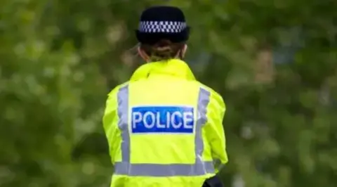 Getty Images The back of a female police officer wearing a yellow reflective jacket and a black hat with bushes in the background