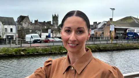 Rachel Duffy stands in front of Newry Canal. She is wearing a tan coloured shirt and has her dark hair pulled back from her face, She is smiling