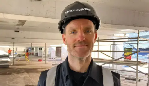 A man stands in a multi-storey car park and smiles, while wearing a black hat with says Capital&Centric. There is construction work taking place behind him.