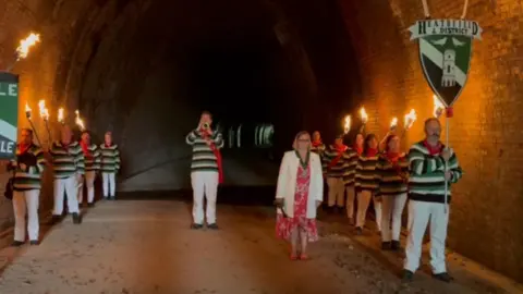 Mick Gosden Lit torches being held in a disused railway tunnel in Heathfield in West Sussex. There is a trumpeter in the middle and people lined up along the edges of the tunnel holding torches and banners.