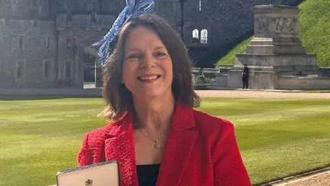 A woman with shoulder-length brown hair with a blue fascinator in it is wearing a red jacket and black top. She is stood in the grounds of an old castle holding a box which contains an MBE medal.