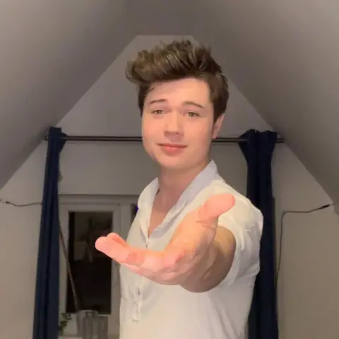 Instagram wearing a white shirt put his hand out of the camera. He stands with a window inside a small room.