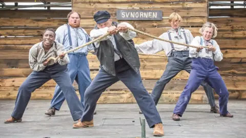 Seed Sedgemoor Five people dressed in suit trousers and shirts are on a stage holding on to a rope.