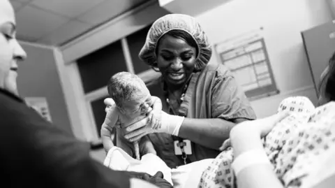 James Clifford Kent A black and white image of a midwife wearing a surgical cap and mask, carefully lifting a crying newborn baby to hand the infant to the mother.