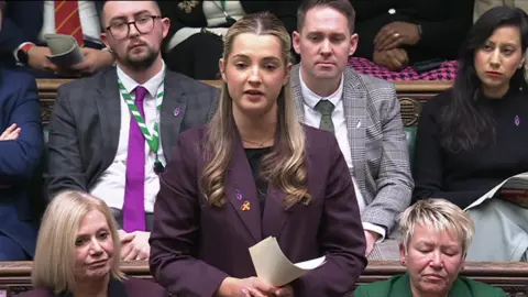 House of Commons Wrighting, talking in the House of Commons, holding a piece of paper. She has long blonde hair, tied back, and is wearing a purple jacket. Other people are sitting around her. 